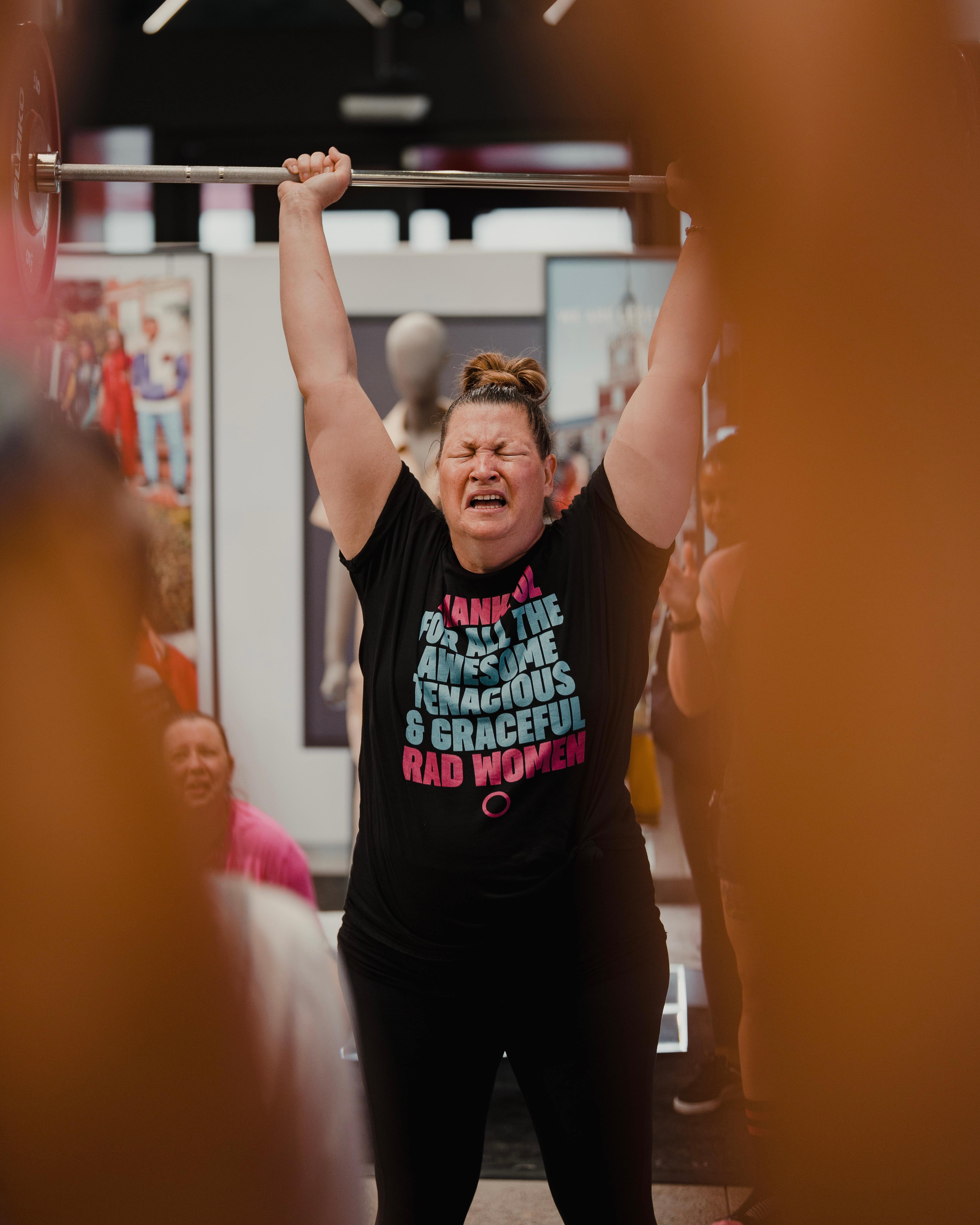 Woman performing an overhead barbell lift wearing a Thankful for RAD Women shirt